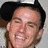 A static image of a person wearing a black cap with white text, smiling broadly against a light background.