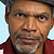 Static pixelated image of a person with brown skin wearing a dark red shirt, light blue background, and visible facial features including eyes, nose, mouth, and beard stubble.