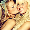 Static image of two blonde women kissing on the cheek with one woman wearing a blue flower accessory in her hair and both having light skin tones.