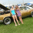 Two figures stand side-by-side in front of a yellow vehicle on grass, one wearing light blue top and shorts, the other purple top and dark shorts.