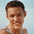 Pixelated close-up of a person with short dark hair, light skin, and a silver chain necklace against a light blue background.