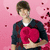 A person in a dark blue jacket holds two red heart-shaped objects against a pink background with scattered hearts.