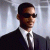 Pixelated image of a person in a dark suit and white shirt, wearing dark sunglasses, facing right against a light gray background.