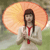 A pixelated figure in traditional attire holds a large orange parasol against a blurred green and brown background.