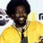 Pixelated image of a person with dark curly hair wearing a yellow shirt and dark tie, smiling against a light background with partial dark shapes.