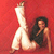 A pixelated image shows a person in a black sleeveless top and white pants against a solid red background, with legs extended upward and one arm resting on the knee.