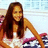 A pixelated image of a person with long brown hair wearing an orange top and white shorts, holding a pink object against a light blue background.