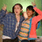 Two people pose with raised arms against a green background; one wears blue jacket, the other striped shirt and pink hoodie.