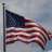 Static pixelated image of a waving American flag on a pole against a light blue sky background.