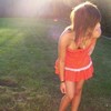 A person in a red strapless dress with white stripes stands on grass, head turned down, backlit by bright light.
