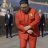 A pixelated figure in a red jumpsuit stands with arms crossed against a blurred background of indistinct shapes and muted colors.