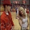 Two people stand on a basketball court; one wears a red outfit and hat, the other a white tank top, with a blurred crowd in the background.
