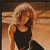A pixelated image of a person with long blonde hair wearing a black sleeveless top, standing against a warm-toned background with horizontal stripes.