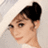 Pixelated image of a person with dark hair styled in an updo, wearing light-colored clothing and a white head covering, positioned against a neutral background.