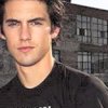 A person with light brown hair wearing a black shirt stands in front of a building with grid-patterned windows.