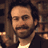 Static pixelated image of a man with dark hair and beard wearing a white shirt under a dark jacket, set against a blurred background with warm brown tones.