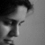 Black-and-white pixelated image of a person with dark hair, wearing a light-colored shirt and dark jacket, holding an object near their face.