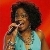 Pixelated static image of a person with dark curly hair singing into a microphone against a red background, wearing a light blue sleeveless top.