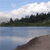 Pixelated landscape with blue sky, white clouds, dark green trees, and a calm body of water reflecting the sky and trees.