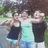 Three people stand closely together outdoors with arms around each other, wearing casual summer clothing against a blurred green and brown background.