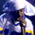 Pixelated image of a figure in dark clothing holding a microphone against a blue and white background with yellow accents.