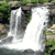 A static pixelated image of a waterfall with white water flowing over dark rocks, surrounded by green foliage and brown earthy terrain.