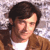 Pixelated close-up of a person with dark brown hair wearing a brown jacket over a white shirt against a light gray background.