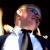 Pixelated static image of a person in formal attire with visible facial features and accessories against dark background.