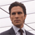 Pixelated portrait of a person with short dark hair wearing a dark suit jacket, white shirt, and dark tie against a white background with gray diagonal lines.