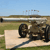 A large artillery piece with a long barrel sits on a grassy field under a clear blue sky, positioned on a concrete base with visible wheels and metal components.
