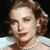 Pixelated close-up of a person with light brown hair styled in soft waves, wearing red lipstick and a white garment against dark background.