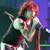 Pixelated image of a person with reddish-brown hair holding a microphone, wearing a green sleeveless top and dark pants against a purple and green background.