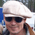 A person wearing a light-colored flat cap and dark sunglasses, with long hair tied back, against an outdoor background.