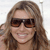 A pixelated close-up of a person with long brown hair wearing dark sunglasses and a light-colored top, smiling with visible teeth against a light background.