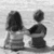 Black-and-white silhouette of two figures sitting side-by-side on a bench, facing left, with a blurred background.