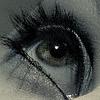A close-up of a human eye with long dark eyelashes, a greenish-brown iris, and a small white reflection in the pupil, set against a blurred light gray background.