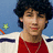 Pixelated image of a person with curly dark hair wearing a red shirt and white jacket with dark stripes, standing against a blue background with a green star and orange shape.