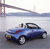 A blue convertible car with a red stripe on the hood is positioned on a road near a body of water, with a bridge structure visible in the background.