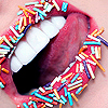 A close-up of a mouth with white teeth and a pink tongue, surrounded by colorful sprinkles on a red surface.
