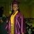 A person in a purple graduation gown and cap with yellow accents stands against a blurred background of seated figures.