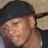 Pixelated close-up of a person wearing a dark cap with red trim, light skin tone, and visible ear.