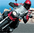 Pixelated image of a character in a red helmet and black outfit riding a motorcycle with visible exhaust smoke against a blue sky background.