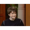 A young person with brown hair in a dark sweater smiles leftward against an indoor background.