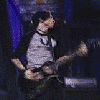 A musician in a gray and black raglan shirt plays electric guitar on stage with blue curtains, moving through performance poses.