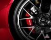 Close-up of a car wheel with red brake caliper and VW logo on the hubcap, showing metallic silver spokes against dark tire and red vehicle body.