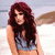 A woman with long red hair in a light blue crop top sits on sand; next to her, a shirtless man kneels in dark jeans and black wristbands.