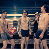 Five shirtless men stand in a row on bleachers; one holds a red-and-yellow object, another wears a cap.