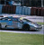 Static image of a blue sports car on a grassy field with a building in the background, rendered in low-resolution pixel art style.