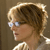 Pixelated profile view of a person with short brown hair wearing light-colored glasses and a light shirt against a brown background.