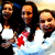 Three people in white tops pose together with a red object held between them against a light background.